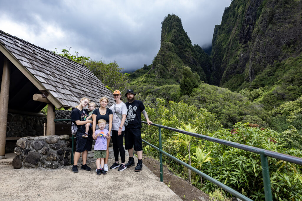 A family of six stands together in a lush green landscape, with mountains in the background and a cloudy sky. They are smiling and enjoying their outdoor adventure near a wooden shelter.