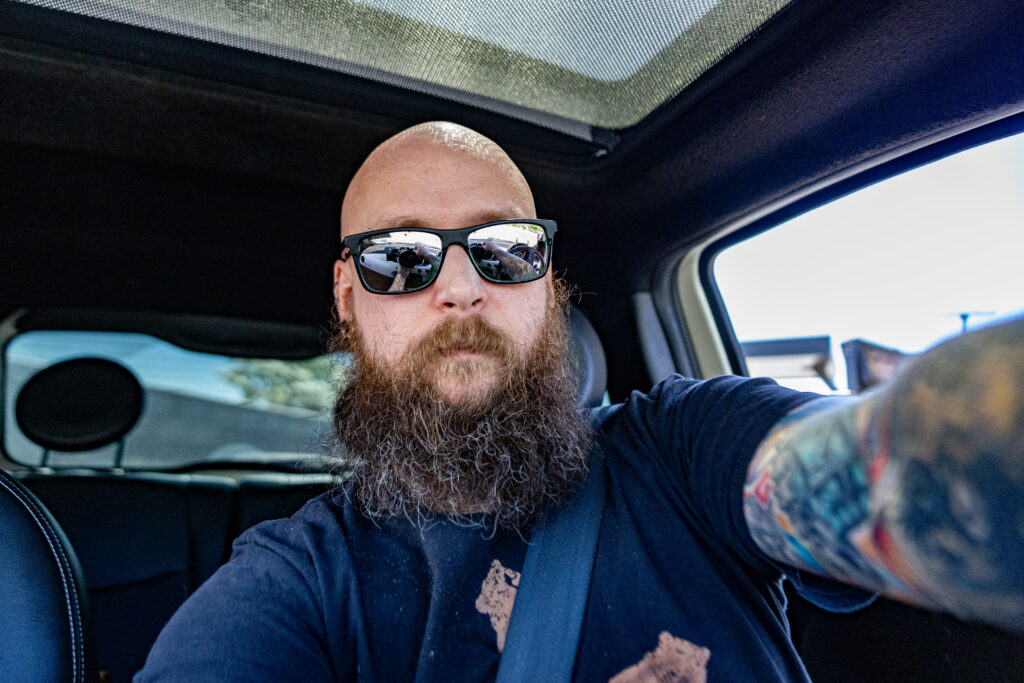 A person with a bald head and a full beard, wearing sunglasses, taking a selfie while sitting in a car.